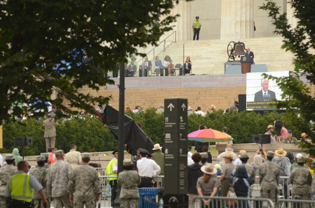 Army personnel and civilians stand listening to a speech given by the president, with some seated on chairs, and a backdrop of bushes, trees, and stairs.
