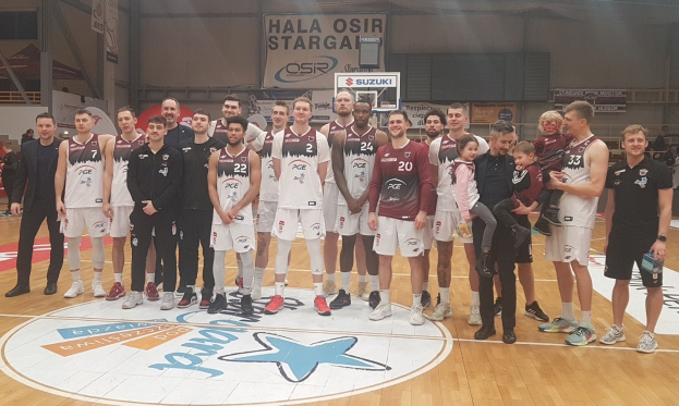 Männergruppe auf einem Basketballfeld mit Kindern und Bannern mit der Aufschrift "Hala Osir Starga" im Hintergrund.
