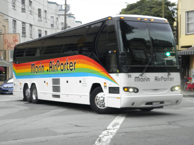 Ein Bus fährt auf einer Straße mit Gebäuden, Bäumen und Polen im Hintergrund.