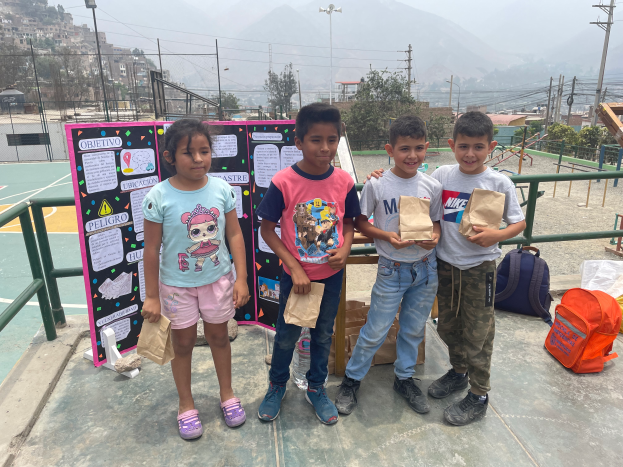 Eine Gruppe von Kindern steht auf einem Basketballfeld, jeder hält einen Papiertüte, mit Tüten und Gegenständen rechts, einer Texttafel links und einem Hintergrund aus Zaun, Pfählen, Straßenlaternen, Bäumen, Gebäuden, Hügeln und Himmel.