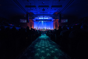 Ein großer Saal voller Menschen auf Stühlen, eine Bühne mit Musikern und eine Decke mit Lichtern und Fernsehgeräten, was auf einen Konzertsaal für die Nationale Convention 2019 hindeutet.