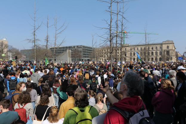Eine große Menge Menschen mit Schildern und Taschen steht vor einem Gebäude mit Fenstern, Bäumen und einem klaren Himmel, was auf eine Klimawandeldemonstration hinweist.