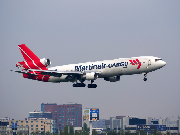 Ein Martinair Cargo Boeing 777-300ER startet vom Frankfurter Flughafen, mit Gebäuden, Bäumen, Polen und Plakaten im Hintergrund und dem Himmel oben.
