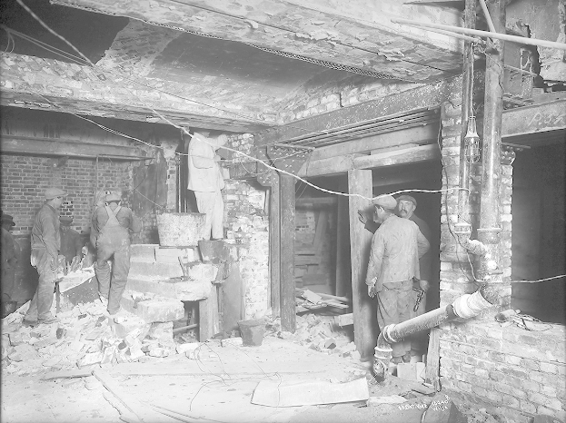 Eine Gruppe von Männern steht in einem durch ein Feuer beschädigten Raum voller Schutt wie Ziegelsteine und Rohre, dargestellt in Schwarz-Weiß.