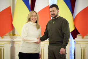 Ein Mann im Anzug und eine Frau in legerer Kleidung geben sich die Hand, lachend, vor einer Flaggenwand, mit einer Wand im Hintergrund.