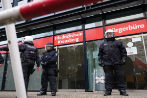 Eine Gruppe von Polizisten in Helmen und Handschuhen steht vor einem Glasgebäude mit einer Stange links und Tafeln im Hintergrund, auf denen "Friedrichshain Kreuzberg" steht und Papiere an den Glaswänden kleben.
