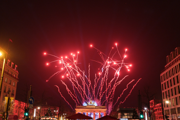 Eine belebte Stadtstraße an einem Neujahrsabend in Berlin mit Gebäuden, Bäumen, Laternen, Ampeln, Schildern, Zelten, Menschen und einem prächtigen Feuerwerk am Himmel.