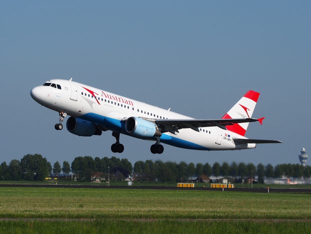 Ein Airbus A320-200 der Austrian Airlines startet von Frankfurt Airport, mit grünem Gras darunter, Bäumen und Gebäuden im Hintergrund und einem Turm auf der rechten Seite, unter einem klaren Himmel.