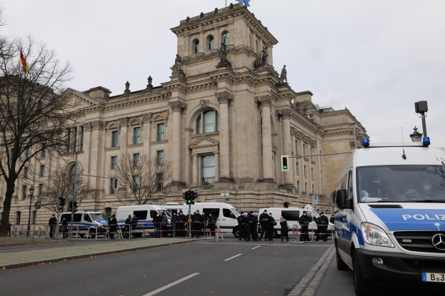Eine Gruppe von Polizisten steht vor dem Reichstaggebäude in Berlin, Deutschland, mit Fahrzeugen, einem Zaun, Verkehrsampeln, Laternenpfählen, Bäumen und Flaggen im Hintergrund, bei klarem Himmel.