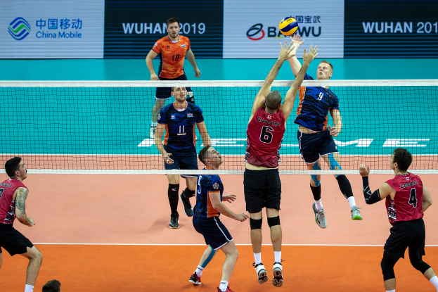 Eine Gruppe von Männern, die Volleyball auf einem Court mit einem Netz und einem Ball in der Luft spielen, mit Tafeln im Hintergrund, die Text und Logos anzeigen, die auf die BWU-Männer-Volleyball-Weltmeisterschaft 2019 hinweisen.