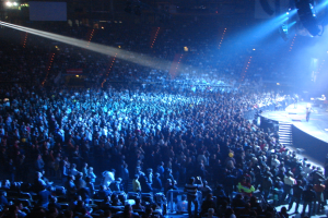 Eine große Menge in einem hell erleuchteten Konzerthaus schaut Musikern auf der Bühne hinter Gittern zu.