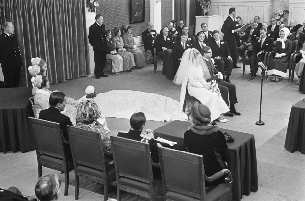 Schwarzes und weißes Foto einer Hochzeitszeremonie mit sitzenden und stehenden Menschen, einem Mann mit Mikrofon und einer Frau mit Blumenstrauß, mit einem Podium mit Mikrofon, Blumenschmuck, einem Vorhang, einem gerahmten Wandfoto und einer Tür im Hintergrund.