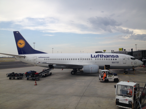 Ein Lufthansa Airbus A320-200 auf dem Frankfurter Flughafen geparkt, mit verschiedenen Bodenfahrzeugen, Wagen, Verkehrskegeln, Pfosten und Texttafeln im Bild, unter einem bewölkten Himmel.