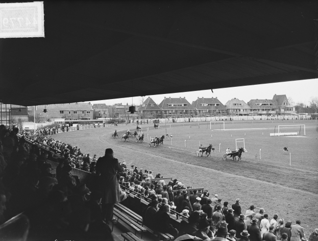 Schwarzes und weißes Foto eines Pferderennens in einer Arena mit Zuschauern auf Bänken und Jockeys, die Pferde auf der Bahn reiten, mit Gebäuden, Bäumen und Pfählen im Hintergrund bei klarem Himmel.