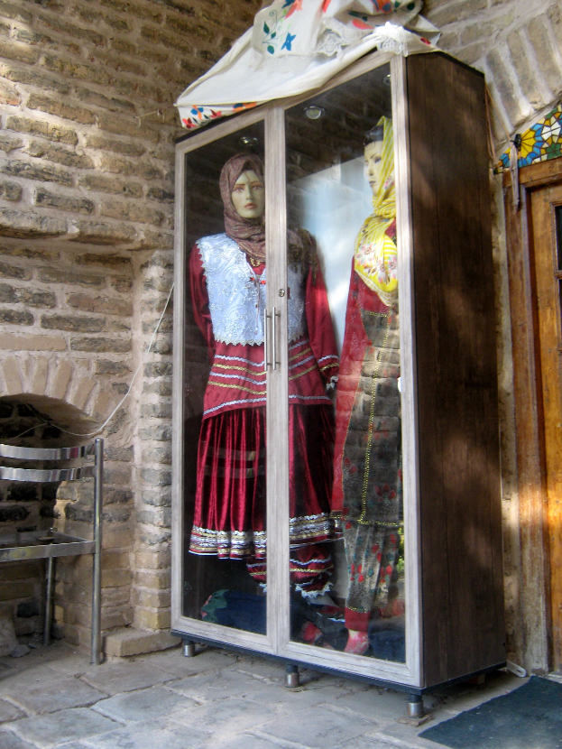 Zwei Schaufensterpuppen in traditioneller türkischer Kleidung sind in einer Glasvitrine mit einem drapierten Tuch, begleitet von einem Stuhl links und einer Tür im Hintergrund, ausgestellt.