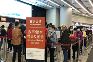Menschen mit Masken, die in einer Reihe vor einer Absperrung mit einer Tafel stehen, auf der "Hong Kong Airport Refunds Due to Covid-19" steht.