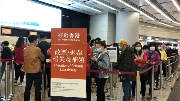 Menschen mit Masken, die in einer Reihe vor einer Absperrung mit einer Tafel stehen, auf der "Hong Kong Airport Refunds Due to Covid-19" steht.