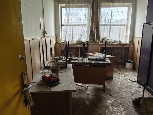 Leerer Raum mit einer Tür auf der linken Seite, einem Schrank auf der rechten Seite und einem Fenster mit Vorhängen im Hintergrund; ein zentraler Tisch hält verstreute Gegenstände, was auf ein verlassenes Büro hindeutet.