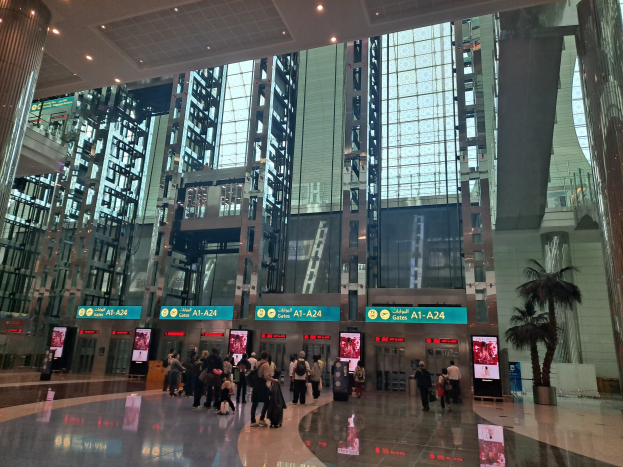 Menschen, die durch eine Flughafenhalle gehen, umgeben von hohen Gebäuden, Bäumen, Texttafeln, Säulen und Deckenleuchten, einige tragen Taschen.