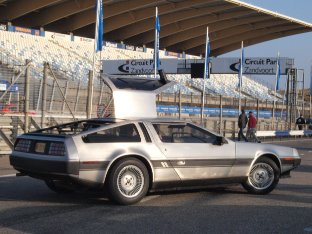 Ein silberner DeLorean DMC-12 fährt auf einer Straße umgeben von Menschen, Fahnen, Schildern, Geländern, Stühlen und einer Hütte, mit Bäumen und einem klaren blauen Himmel im Hintergrund.