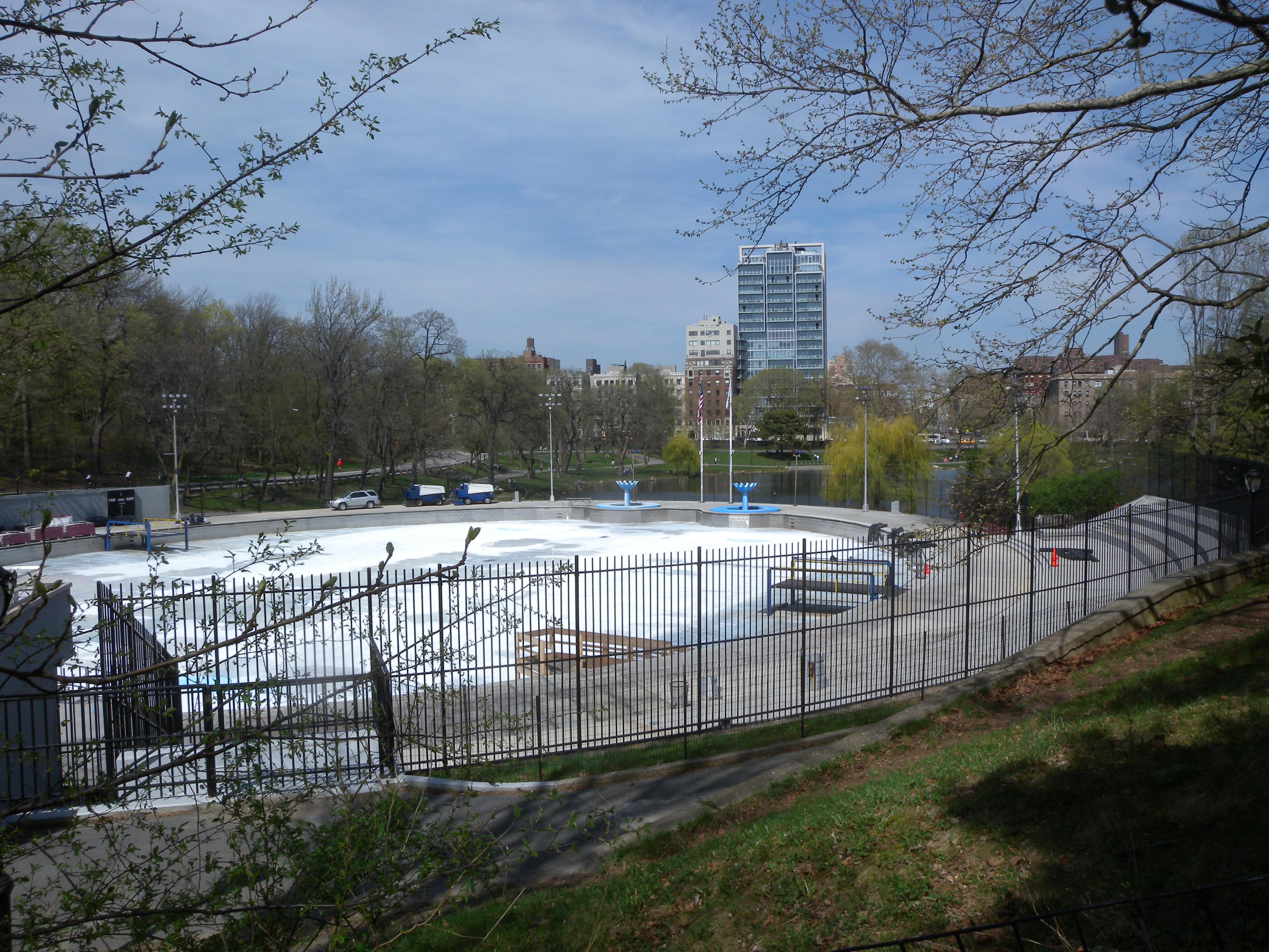 Eine Eisbahn in einem Park, umgeben von einem Metallzaun, Gras, Pflanzen, Bäumen, Fahrzeugen, Straßenlaternen, einem Gewässer, Gebäuden mit Fenstern und einem bewölkten Himmel.