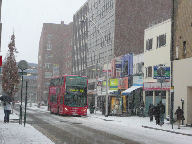 Ein roter Doppeldeckerbus fährt eine schneebedeckte Straße mit hohen Gebäuden, Menschen mit Regenschirmen, Laternen, Texttafeln und Bäumen entlang.