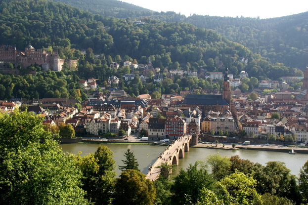 Eine malerische Ansicht von Heidelberg, Deutschland, mit einer Brücke über einen Fluss, fahrenden Fahrzeugen, Gebäuden mit Fenstern, Bäumen, fernen Hügeln und einem klaren blauen Himmel.