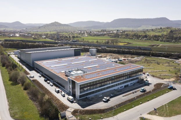Luftaufnahme eines großen Industriegebäudes mit Solarmodulen auf dem Dach, umgeben von Fahrzeugen, Bäumen, Gras, Strommasten und umgeben von Hügeln und einem klaren blauen Himmel.