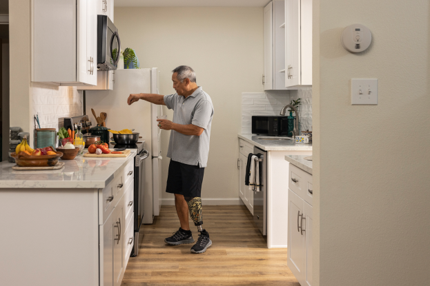 Ein Mann steht in einer Küche mit weißen Schränken und hält ein Glas in der Hand, links auf der Arbeitsplatte stehen Obst und Gemüse, Schüsseln, Löffel und andere Gegenstände, im Hintergrund verschiedene Geräte und Gegenstände.