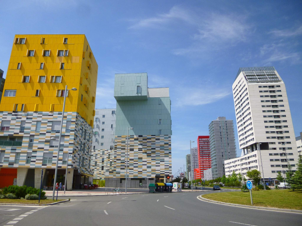 Eine Straße in einer Stadt mit hohen Gebäuden im Hintergrund, Straßenmasten, Straßenlaternen, Kraftfahrzeugen, Infotafeln, Büschen, Bäumen und einem bewölkten Himmel, mit den einzigartig gestalteten Gebäuden der Universität Rotterdam.