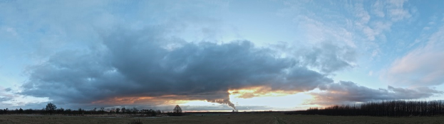 Ein bewölkter Himmel mit einem Fabrikschacht, aus dem Rauch aufsteigt, und ein paar Bäumen und Pflanzen auf der rechten Seite.