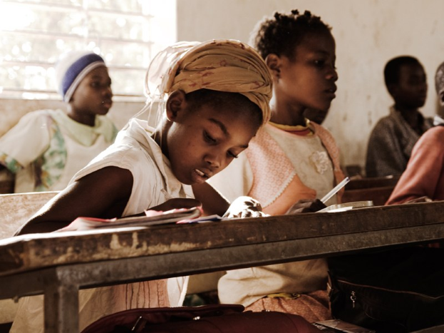 Kinder sitzen an Tischen in einem Klassenzimmer mit Büchern und Taschen vor sich, mit einer Wand und einem Fenster im Hintergrund.