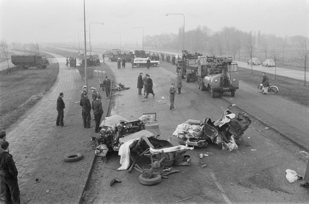Schwarz-weiß-Szene eines Autounfalls am Straßenrand mit mehreren Fahrzeugen und einer Gruppe von Menschen drumherum, Laternenmasten und Bäume im Hintergrund.