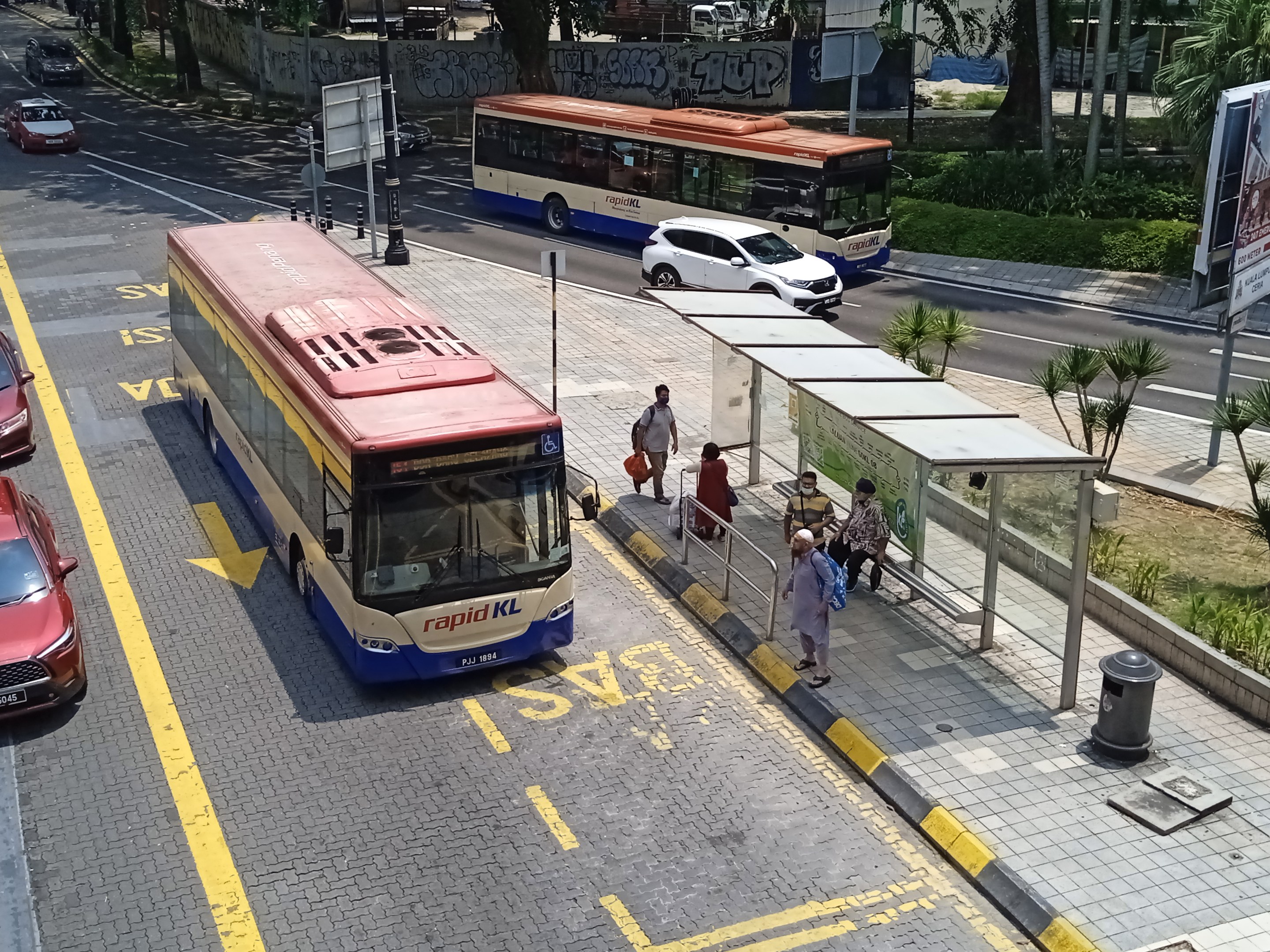 Stadtstraße mit fahrenden Autos und Bussen, ein Bushaltestelle mit Fußgängern auf dem Gehweg, umgeben von Pfählen, Schildern, Bäumen, Gras und Gebäuden.
