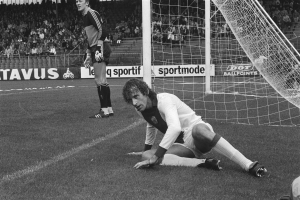 Ein Schwarz-Weiß-Foto eines Fußballspielers, der vor einem Tor auf dem Boden kniet, umgeben von einer Gruppe von Menschen auf einem Rasenfeld, mit einem Zaun, einer Fahne, Pfosten, einer Treppe und einem Dach mit Deckenlampen im Hintergrund.