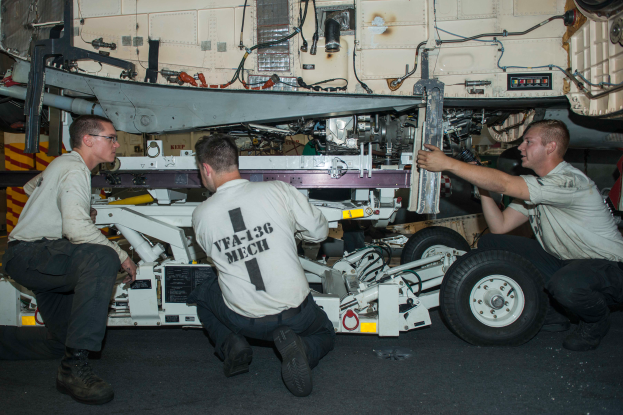 Drei Männer in Uniformen arbeiten an einem Flugzeug in einer Halle, umgeben von Werkzeugen und Ausrüstung, scheinbar Reparaturen durchführend.