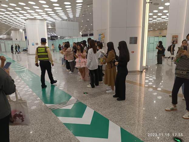 Eine Gruppe von Menschen steht in einer Schlange an einem Flughafen-Sicherheitscheck, einige halten Taschen und Mobiltelefone, mit Säulen im Hintergrund und Lampen an der Decke und einem Wasserzeichen am unteren Rand des Bildes.