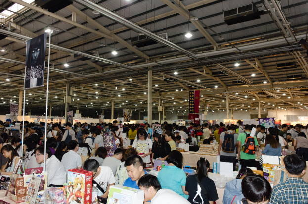 Eine große Gruppe von Menschen versammelte sich um Tische, die mit Büchern, Kartons und anderen Gegenständen gefüllt waren, auf einer Buchmesse, mit Bannern und Lichtern darüber.