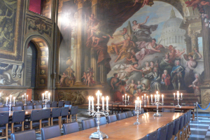 Tische und Stühle sind in einem Raum angeordnet, mit Kerzen auf Ständern auf den Tischen. Im Hintergrund ist eine Wand mit einem Bogen und mehreren paintings zu sehen.