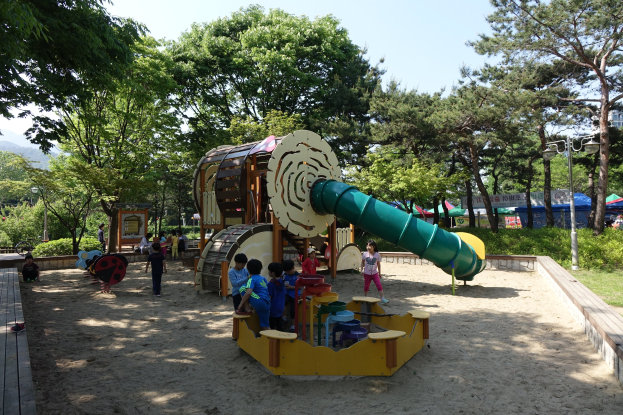 Kinder auf einem Spielplatz mit einer Rutsche, Sand und Bäumen im Hintergrund, während andere auf einer hölzernen Plattform sitzen; Zelte, Laternenmasten und ein klarer blauer Himmel sind zu sehen.