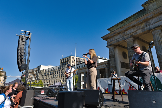 Eine Gruppe von Menschen auf der Bühne vor dem Brandenburger Tor in Berlin, Deutschland, die Musikinstrumente spielen und in Mikrofone singen, mit Lautsprechern und anderen Gegenständen drumherum und Gebäuden, Bäumen, Pfählen, Flaggen und einem klaren blauen Himmel im Hintergrund.