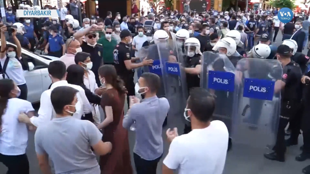 Eine Gruppe von Menschen vor einer Reihe von Polizisten mit Helmen und Schilden, mit einem geparkten Auto links und einer Person mit einer Kamera rechts, vor Gebäuden und Bäumen in Ankara, Türkei.