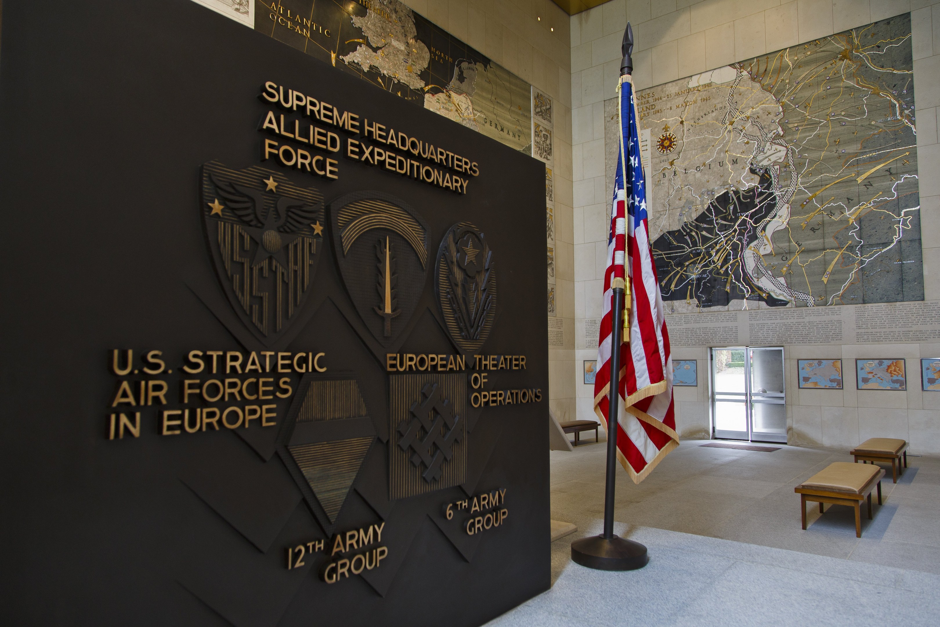 Eingang zum U.S. Strategic Air Forces Headquarters in Europa, mit einer amerikanischen Flagge, einer Wand mit Text und Logos, Bänken, einer Tafel, Fotorahmen, beleuchteter Decke und einer Tür im Hintergrund.