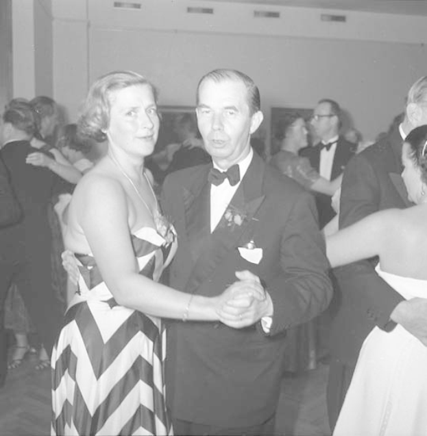 Zwei Tänzer, ein Mann in einem weißen T-Shirt und blauen Hosen und eine Frau in weißen Hosen und einer grünen Oberteil, umarmen und bewegen sich anmutig auf einer hell erleuchteten Bühne mit einem bunten Hintergrund.