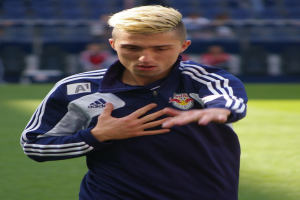 Ein junger Mann in einem blauen T-Shirt steht auf einem Fußballfeld und streckt die Arme aus, wobei er entschlossen wirkt.