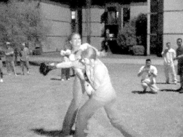 Zwei Personen boxen in der Mitte eines Schwarz-Weiß-Bildes, im Hintergrund sind ein Gebäude und einige Zuschauer zu sehen.