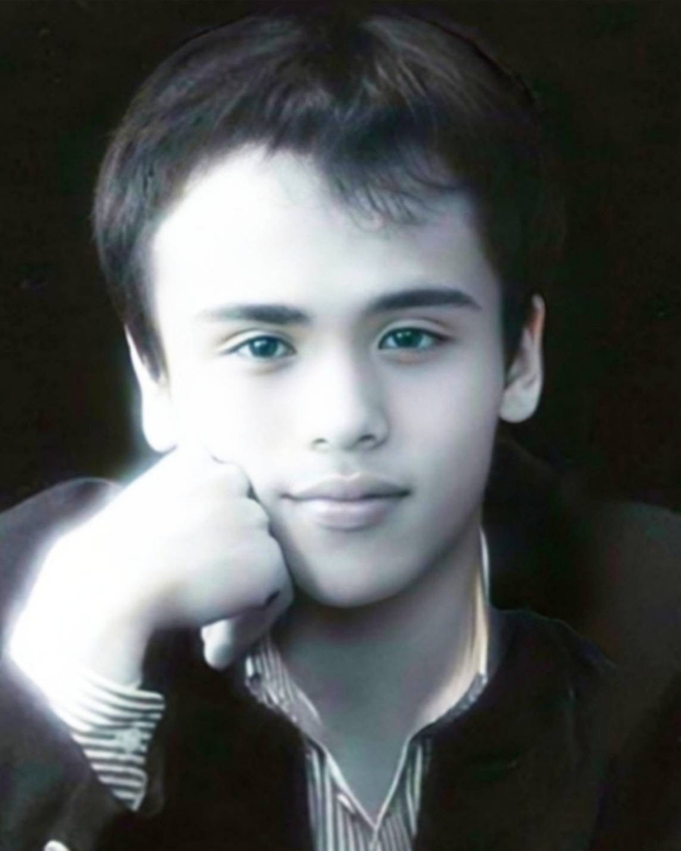 Ein selbstbewusster junger Mann in einer schwarzen Jacke posiert vor einem dunklen Hintergrund mit entschlossener Miene, leuchtenden Augen und ordentlich frisiertem Haar.