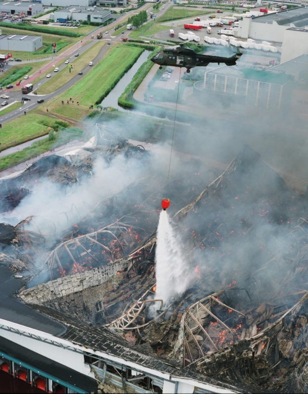 Ein Hubschrauber fliegt über ein großes, durch ein Feuer zerstörtes Gebäude mit Rauch, der daraus aufsteigt, vor einer Kulisse aus Gebäuden, Fahrzeugen, Bäumen und Text am unteren Rand.