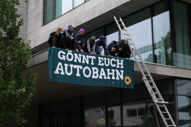 Eine Gruppe von Menschen auf einem Gebäude, die ein Banner halten, mit einem Baum links und Glasfenstern im Hintergrund, die scheinbar für die deutsche Regierung protestieren.