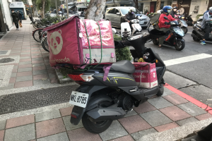 Ein Kickboard steht auf der Seite einer Straße neben einem Gebäude, mit einem Auto, einem Fahrrad und einigen Menschen in der Nähe und Bäumen, Gebäuden und Schildern mit Wörtern im Hintergrund.